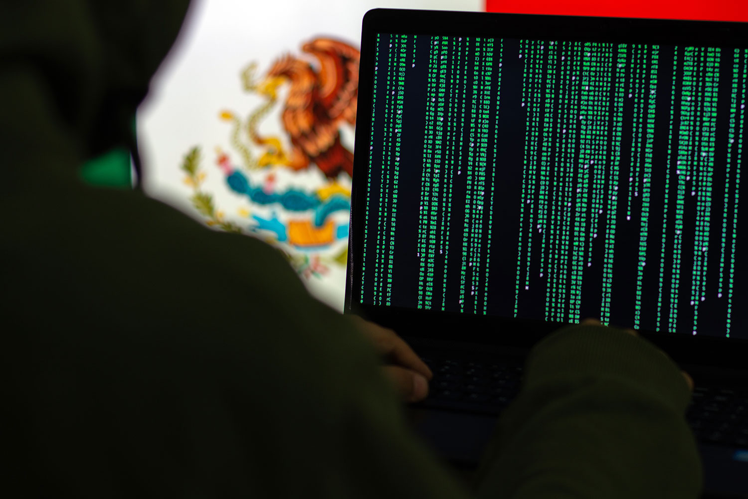un hacker con una computadora portátil y la bandera de México al fondo