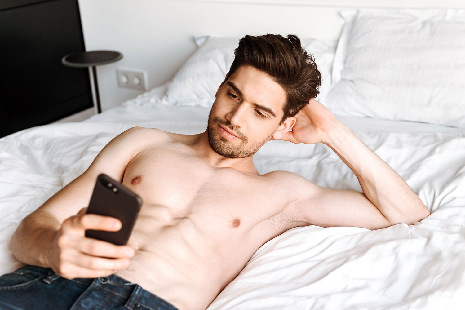 un hombre con el torso desnudo mira su teléfono móvil tumbado en la cama
