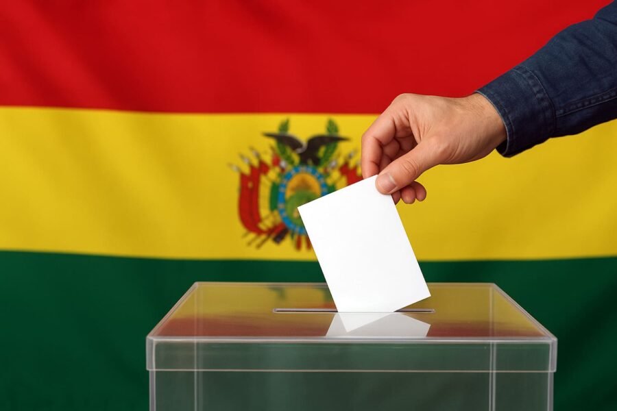 una mano introduce una papeleta en una urna electoral con la bandera de Bolivia en el fondo