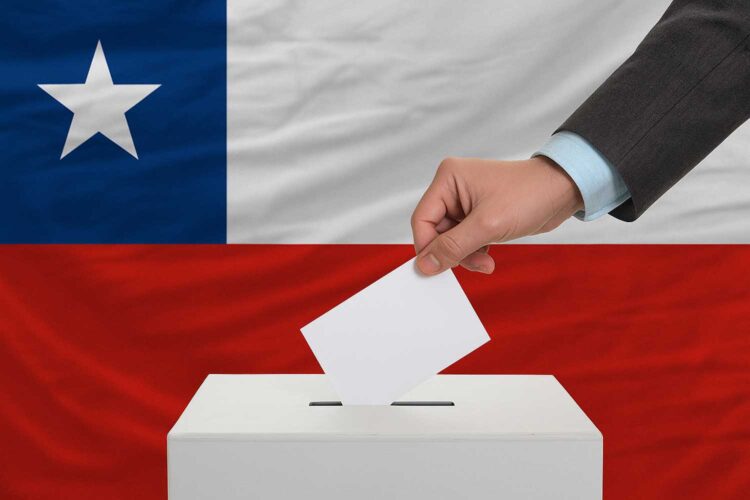 una mano introduce un sobre en una urna electoral con la bandera de Chile al fondo
