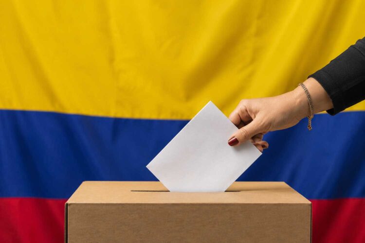 una mujer introduce una papeleta en una urna con la bandera de Colombia al fondo